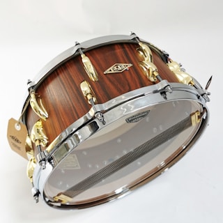 A beautifully crafted snare drum with a wooden finish and metal hardware. The shell displays a rich wood grain texture, complemented by polished chrome and gold hardware, including lugs and hoops. A transparent drumhead allows visibility of the internal components, such as the snare wires.