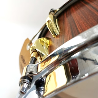 A close-up view of a drum with polished metal hardware and a wooden finish. The shiny gold and chrome components create a contrast with the wood, highlighting the craftsmanship and design details of the instrument.