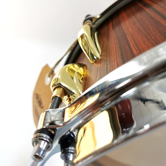 A close-up view of a drum with polished metal hardware and a wooden finish. The shiny gold and chrome components create a contrast with the wood, highlighting the craftsmanship and design details of the instrument.