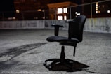 An office chair is placed outdoors on a rough, cracked surface, likely a rooftop or parking area, surrounded by a low barrier fence. The scene is illuminated by surrounding city lights at night, casting soft shadows around the chair.