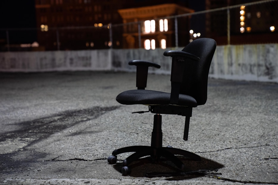 An office chair is placed outdoors on a rough, cracked surface, likely a rooftop or parking area, surrounded by a low barrier fence. The scene is illuminated by surrounding city lights at night, casting soft shadows around the chair.