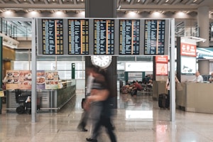 A busy airport terminal with a large electronic flight information board displaying numerous flights and times. Below the board, passengers are moving briskly across the polished floor, some carrying luggage. A clock is visible underneath the flight display. Various signs and advertisements are present, including images of food and a counter where people are seated.