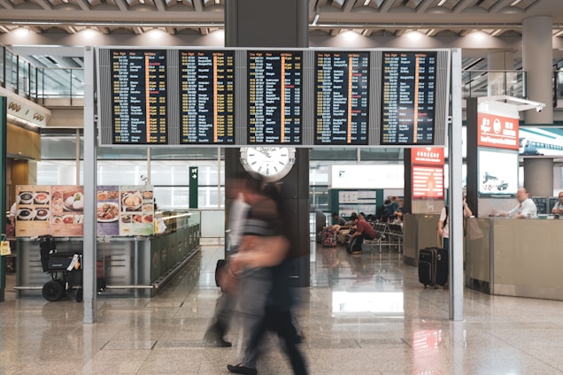 A busy airport terminal with a large electronic flight information board displaying numerous flights and times. Below the board, passengers are moving briskly across the polished floor, some carrying luggage. A clock is visible underneath the flight display. Various signs and advertisements are present, including images of food and a counter where people are seated.