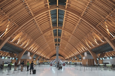 Modern airport terminal featuring an expansive vaulted ceiling constructed from wooden beams. The interior is spacious with check-in counters along one side and several people with luggage. The floors are shiny and the lighting creates a warm ambiance.