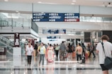 A bustling airport terminal features travelers walking in various directions. Many people are carrying luggage, and others are engaged in conversation. Overhead signs with directions in multiple languages are visible, as well as escalators to the side.