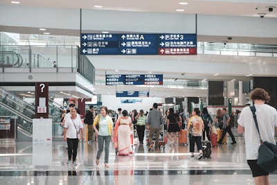 A bustling airport terminal features travelers walking in various directions. Many people are carrying luggage, and others are engaged in conversation. Overhead signs with directions in multiple languages are visible, as well as escalators to the side.