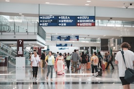 A bustling airport terminal features travelers walking in various directions. Many people are carrying luggage, and others are engaged in conversation. Overhead signs with directions in multiple languages are visible, as well as escalators to the side.