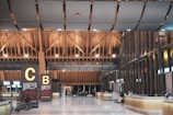 The scene is of an airport terminal with a high, intricate wooden ceiling. The building architecture includes wooden beams and modern design elements. Near the center are two large signs with the letters 'C' and 'B'. The area has shiny white floors and multiple check-in counters or help desks lined up along the right side. There are a few people scattered around sitting and going about their routines, and some wheelchairs are visible next to the signs.