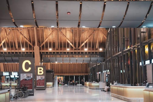The scene is of an airport terminal with a high, intricate wooden ceiling. The building architecture includes wooden beams and modern design elements. Near the center are two large signs with the letters 'C' and 'B'. The area has shiny white floors and multiple check-in counters or help desks lined up along the right side. There are a few people scattered around sitting and going about their routines, and some wheelchairs are visible next to the signs.