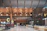 The scene is of an airport terminal with a high, intricate wooden ceiling. The building architecture includes wooden beams and modern design elements. Near the center are two large signs with the letters 'C' and 'B'. The area has shiny white floors and multiple check-in counters or help desks lined up along the right side. There are a few people scattered around sitting and going about their routines, and some wheelchairs are visible next to the signs.