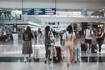 A busy airport terminal with several travelers walking and wheeling suitcases. People are moving in different directions, and overhead signs in multiple languages guide navigation. There are glass railings, escalators, and a high ceiling with bright lighting.