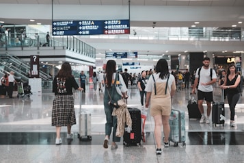 A busy airport terminal with several travelers walking and wheeling suitcases. People are moving in different directions, and overhead signs in multiple languages guide navigation. There are glass railings, escalators, and a high ceiling with bright lighting.