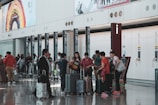 A busy airport terminal features a group of people with luggage near a row of check-in counters. Large advertisements and informational signs are visible above the counters. The floor is reflective, and people appear to be conversing and moving about with their suitcases.