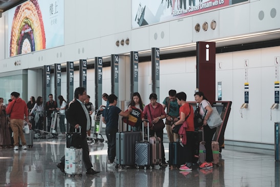 A busy airport terminal features a group of people with luggage near a row of check-in counters. Large advertisements and informational signs are visible above the counters. The floor is reflective, and people appear to be conversing and moving about with their suitcases.