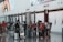 A busy airport terminal features a group of people with luggage near a row of check-in counters. Large advertisements and informational signs are visible above the counters. The floor is reflective, and people appear to be conversing and moving about with their suitcases.