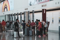 A busy airport terminal features a group of people with luggage near a row of check-in counters. Large advertisements and informational signs are visible above the counters. The floor is reflective, and people appear to be conversing and moving about with their suitcases.