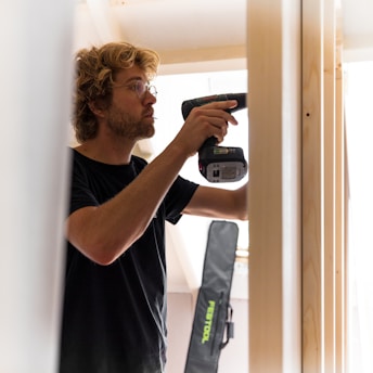 A cheerful woman using a cordless drill while assembling a wooden shelf in a bright, modern farmhouse-style room.