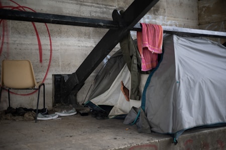 A tent is set up under a concrete structure, supported by large metal beams. A chair with a few pairs of shoes and a pair of gloves are placed to the left of the tent. A colorful towel and some clothing are draped over the metal beam above the tent. One bare foot is visible, sticking out from the tent flap.