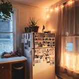 A cozy kitchen corner features a refrigerator covered with various photos and notes, surrounded by warm string lights and a plant on top. A countertop with vegetables and a window with sheer curtains add to the homey atmosphere.