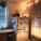A cozy kitchen corner features a refrigerator covered with various photos and notes, surrounded by warm string lights and a plant on top. A countertop with vegetables and a window with sheer curtains add to the homey atmosphere.