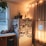 A cozy kitchen corner features a refrigerator covered with various photos and notes, surrounded by warm string lights and a plant on top. A countertop with vegetables and a window with sheer curtains add to the homey atmosphere.