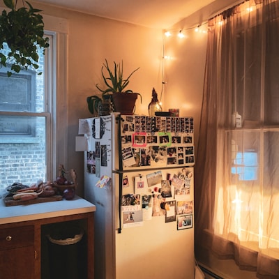 A cozy kitchen corner features a refrigerator covered with various photos and notes, surrounded by warm string lights and a plant on top. A countertop with vegetables and a window with sheer curtains add to the homey atmosphere.