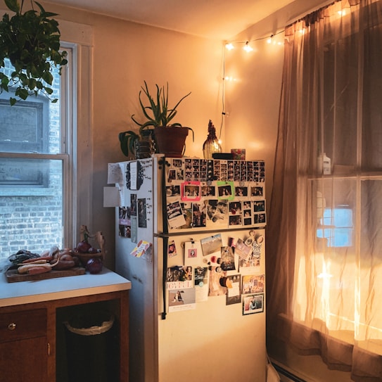 A cozy kitchen corner features a refrigerator covered with various photos and notes, surrounded by warm string lights and a plant on top. A countertop with vegetables and a window with sheer curtains add to the homey atmosphere.