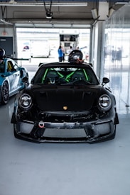 A sleek black sports car parked inside a garage, with a racing helmet placed on its roof. The car has striking green roll bars visible through the windshield. In the background, a few people are standing outside the garage, near other vehicles and equipment.