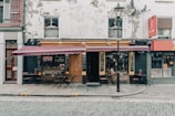 A quaint café with a weathered exterior sits on a cobblestone street. The building features a large open sign, chalkboard menus, and tables with chairs outside under a red awning. Various potted plants add a touch of greenery to the sidewalk seating area.