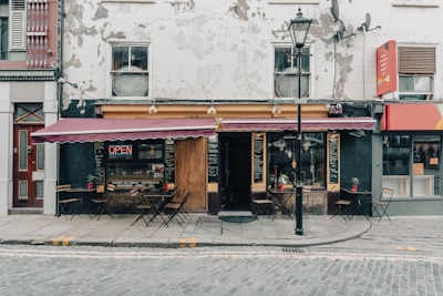 A quaint café with a weathered exterior sits on a cobblestone street. The building features a large open sign, chalkboard menus, and tables with chairs outside under a red awning. Various potted plants add a touch of greenery to the sidewalk seating area.