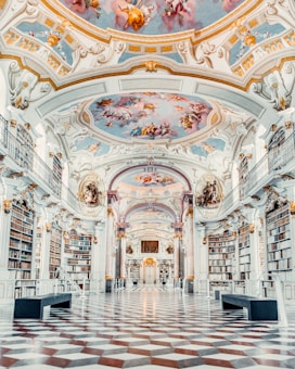 A grand library hall with ornate architecture, featuring towering bookshelves filled with books. The ceiling is elaborately painted with classical frescoes depicting mythological scenes. The floor is patterned with a black and white diamond design, and there are benches for seating.