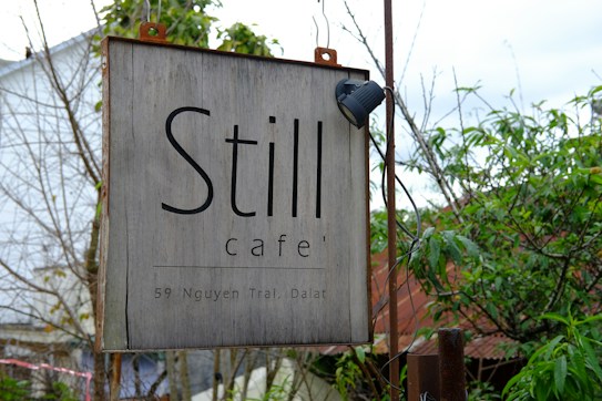 A rustic wooden sign for 'Still Caf&eacute;' is hanging outdoors, featuring a simple font with the address '59 Nguyen Trai, Dalat' below. The sign is framed by greenery and partially visible structures, suggesting a natural, serene setting.