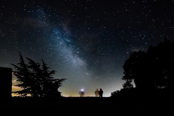 A starry night sky with the Milky Way visible above a silhouetted landscape, featuring trees and two people standing next to a camera on a tripod.