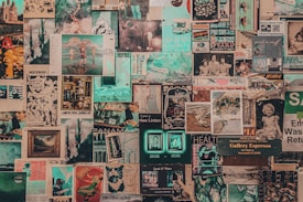 A collage of vintage and contemporary posters and photographs covers a wall. The images include printed art, illustrations, photographs, magazine cut-outs, and handwritten notes. Elements appear layered and overlapping, creating a textured and chaotic visual effect.