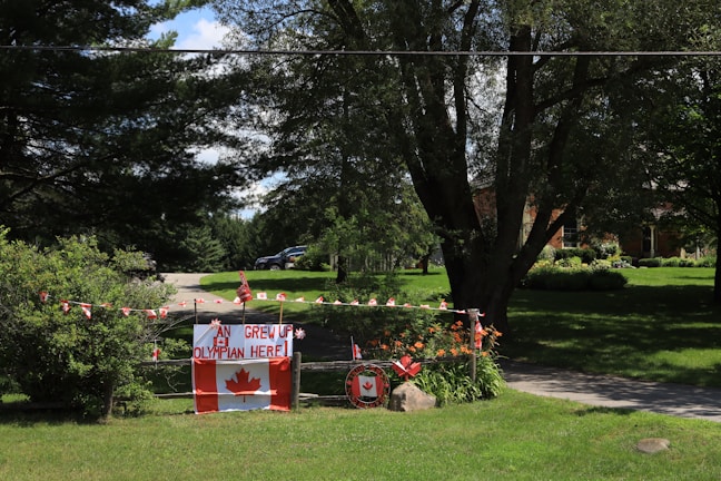 A rural setting features a driveway lined with Canadian flags and a sign that says 'An Olympian grew up here!' adorned with a large Canadian flag and decorative elements. Trees and a grassy lawn surround the area, and there is a brick house partially visible in the background.