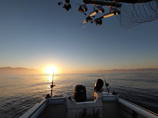 A serene ocean view with the sun setting near the horizon, illuminating the calm water with a warm glow. Two fishing rods are positioned at the back of a boat, and the sky displays a gradient from bright yellow to deep blue. Silhouetted mountains can be seen in the distance, adding to the tranquil atmosphere.