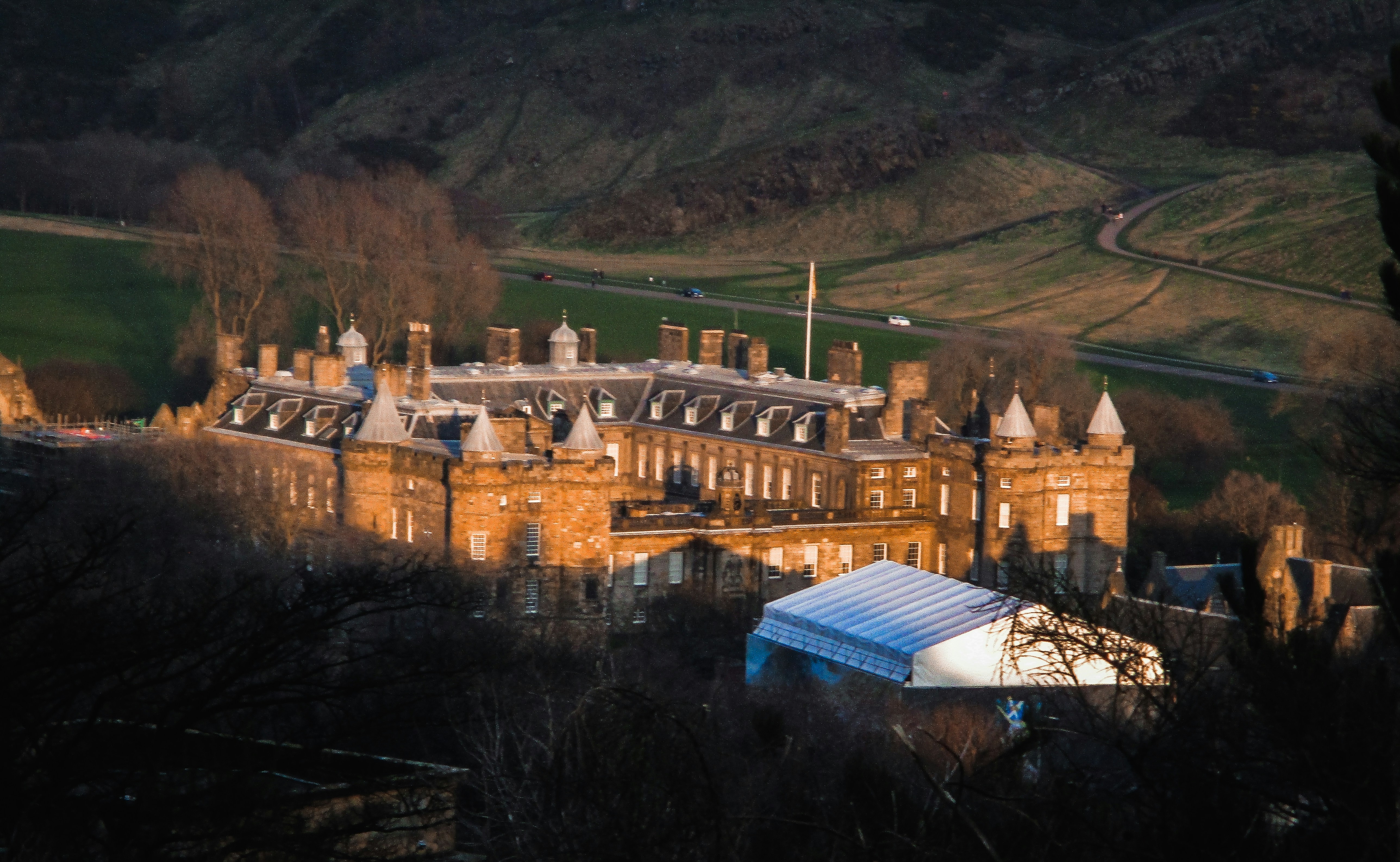 Castle-like stone buildings sit in a valley, bathed in warm dusk light. A white tent-like canopy sits in the foreground with a winding road and bare trees framing the scene.