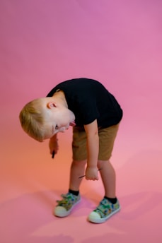 A young child with blonde hair is bending forward playfully while holding two small sticks. The child is wearing a black t-shirt, brown shorts, and colorful sneakers with a vibrant pattern. The background is a soft gradient of pink and orange.
