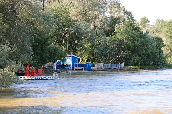 A serene lakeside scene with dense green foliage in the background and a small blue building situated near the water. There is a dock extending into the water with several red chairs placed on it. The water has gentle waves and reflects some of the greenery around it.