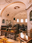An ornate library with a high, decorative arched ceiling featuring inscriptions and a central emblem. Bookshelves line the walls with numerous books organized neatly. A mezzanine level with wooden railings and metal frames circles the library, offering additional seating and tables for reading.