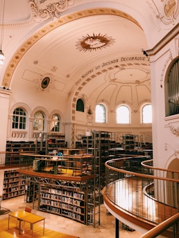 An ornate library with a high, decorative arched ceiling featuring inscriptions and a central emblem. Bookshelves line the walls with numerous books organized neatly. A mezzanine level with wooden railings and metal frames circles the library, offering additional seating and tables for reading.