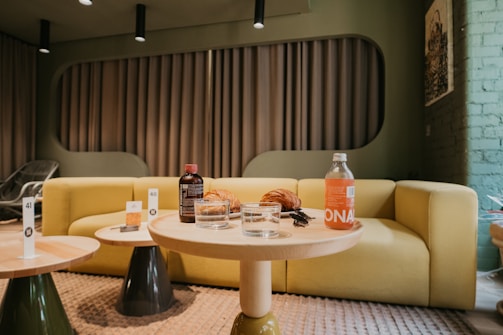 A cozy seating area featuring a yellow sofa against a modern backdrop with geometric patterns. On a small round wooden table in the foreground are two clear glasses, a croissant, a dark brown bottle, and a clear bottle with an orange label. The environment appears relaxed and stylish with a mix of natural tones and subtle lighting.