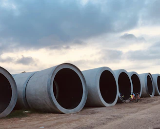 Large cylindrical concrete pipes are laid out in a row along a dirt pathway. A couple of bicycles are placed next to the pipes, and there are people near them. Above, the sky is partly cloudy, creating a mix of light and shadow over the scene.