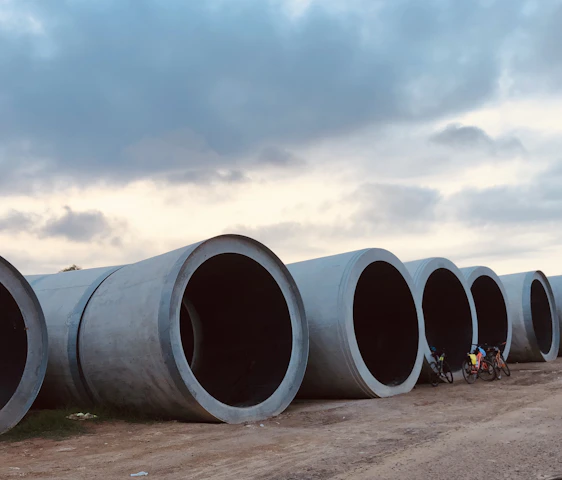 Large cylindrical concrete pipes are laid out in a row along a dirt pathway. A couple of bicycles are placed next to the pipes, and there are people near them. Above, the sky is partly cloudy, creating a mix of light and shadow over the scene.