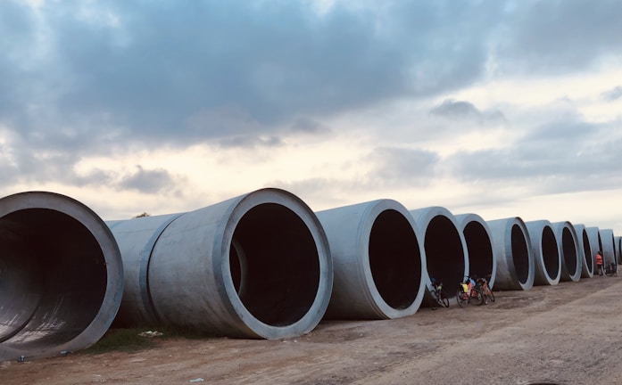 Large cylindrical concrete pipes are laid out in a row along a dirt pathway. A couple of bicycles are placed next to the pipes, and there are people near them. Above, the sky is partly cloudy, creating a mix of light and shadow over the scene.