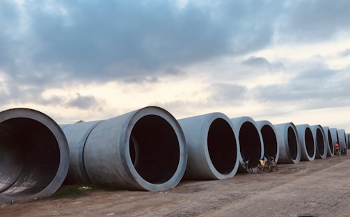 Large cylindrical concrete pipes are laid out in a row along a dirt pathway. A couple of bicycles are placed next to the pipes, and there are people near them. Above, the sky is partly cloudy, creating a mix of light and shadow over the scene.