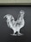 A monochrome illustration of a rooster is depicted on a dark surface. The drawing features intricate line work, highlighting the rooster's feathers and form against a flat backdrop.