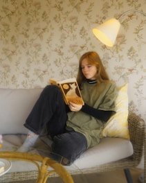 A person with long hair is sitting on a sofa, absorbed in reading a book titled 'Dune'. The room features a floral-patterned wallpaper, and a soft light from a nearby lamp creates a cozy atmosphere.