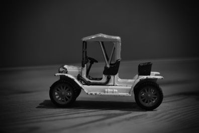 A monochrome image of a miniature vintage car toy with detailed features like large wheels, a canopy top, and an open seating area. The toy is positioned on a wooden surface, with a blurred background that enhances its distinct outlines and features.