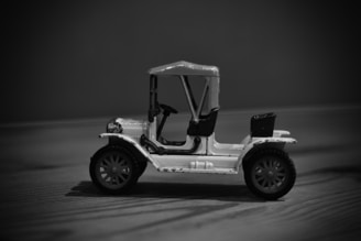 A monochrome image of a miniature vintage car toy with detailed features like large wheels, a canopy top, and an open seating area. The toy is positioned on a wooden surface, with a blurred background that enhances its distinct outlines and features.